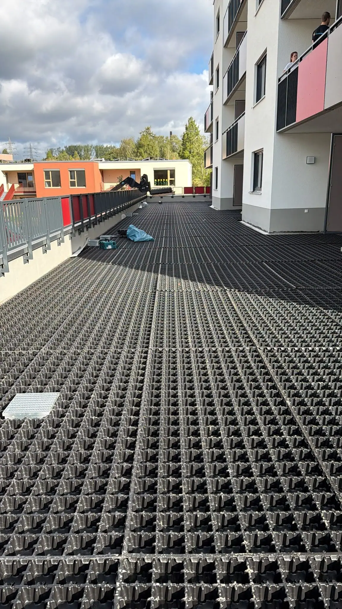Großflächig verlegte Dränelemente mit Kontrollschacht vor einem Mehrfamilienhaus mit roten Balkonverkleidungen