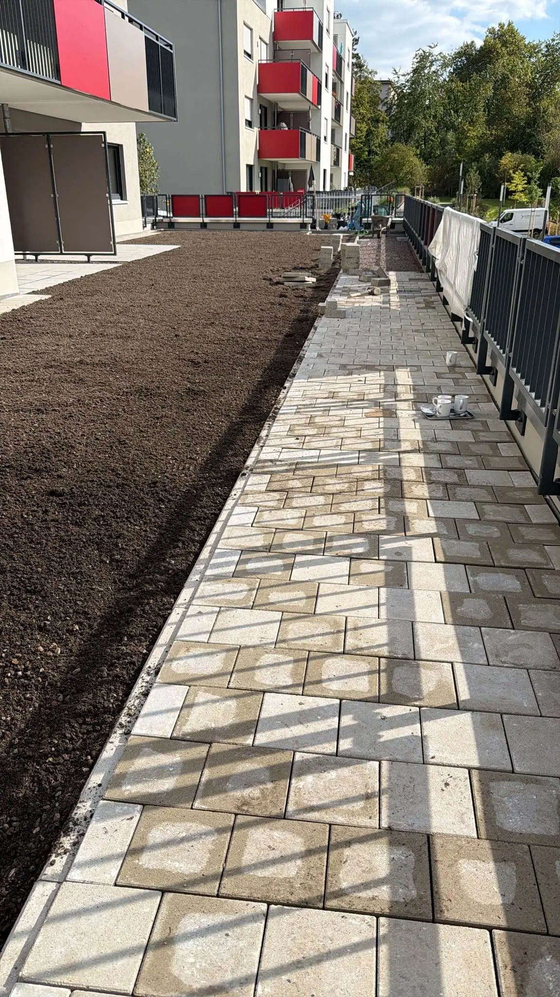Neuer Pflasterweg aus hellem Betonstein entlang eines Geländers, daneben die fertig verteilte Vegetationsschicht vor einer Wohnanlage