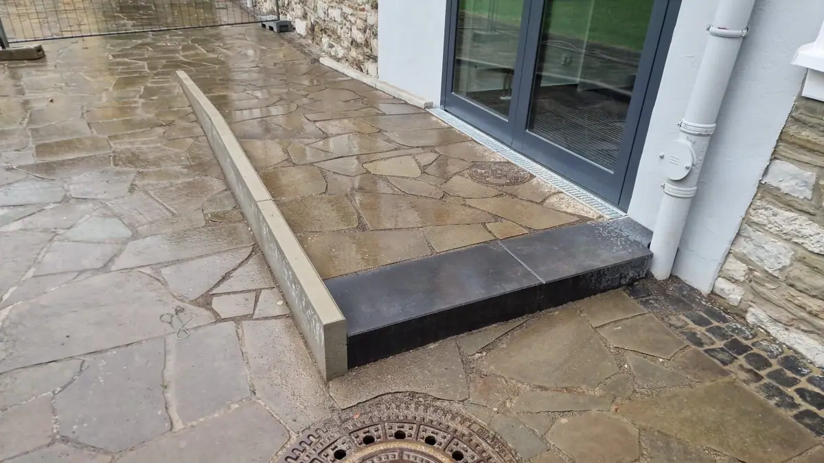Fläche aus Polygonalplatten aus Naturstein vor einer Glastür, eingefasst mit Betonborden und einer anthrazitfarbenen Blockstufe