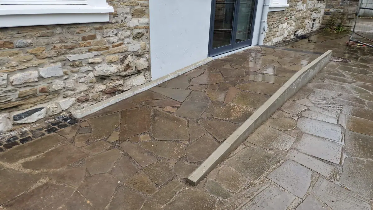 Polygonalplatten aus Naturstein vor der Glastür eines Gebäudes mit Bruchsteinmauerwerk, seitlich eingefasst mit einem Betonbord