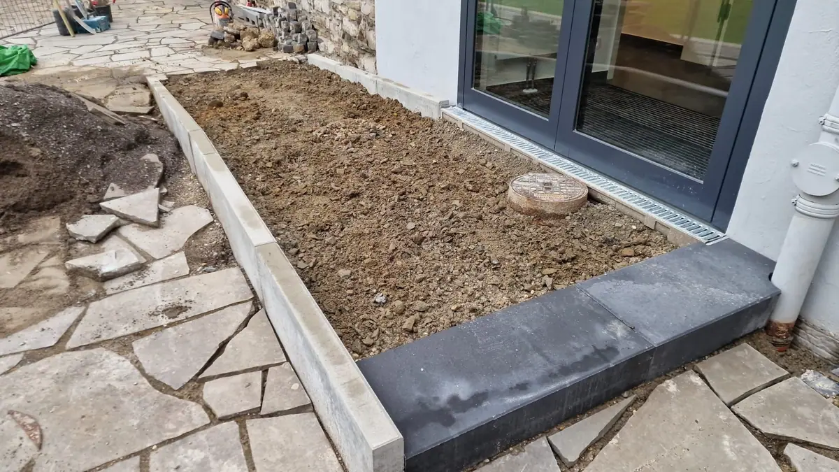 Vorbereiteter Unterbau innerhalb der neuen Einfassung aus Betonborden, mit Blockstufe und Schacht vor einer Glastür
