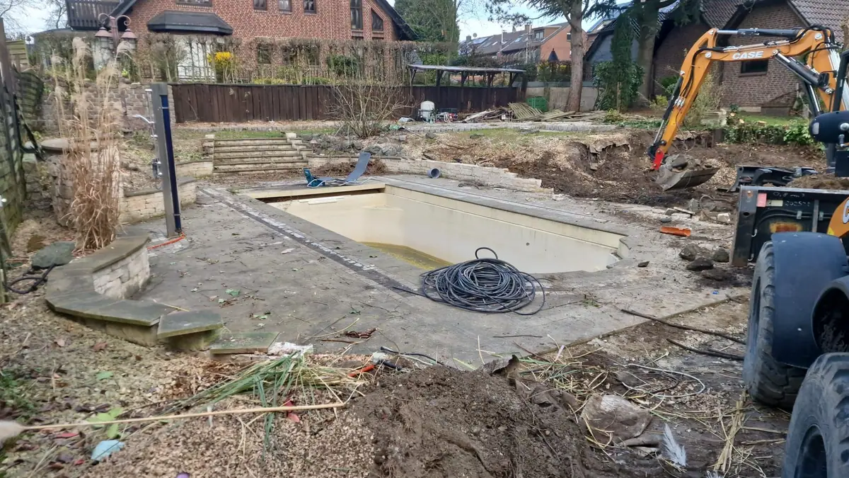 Alter Poolbereich mit verwitterten Platten und verwildertem Garten zu Beginn der Erdarbeiten mit Bagger und Radlader