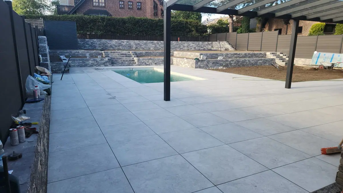 Neue Terrasse aus hellgrauen Großformatplatten rund um einen Pool, dahinter Mauern und Treppen aus grauen Mauersteinen