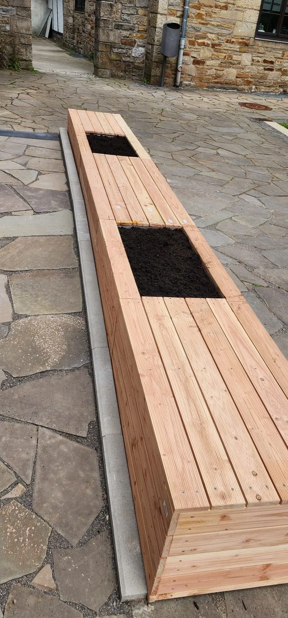 Sitzbank aus Holz mit zwei befüllten Pflanzkästen auf einem Platz aus Polygonalplatten vor Bruchsteinmauern