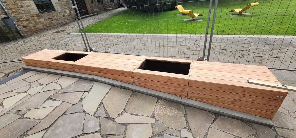 Lange Sitzbank aus Holz mit zwei Pflanzkästen entlang einer Betoneinfassung, darauf eine Wasserwaage zum Ausrichten