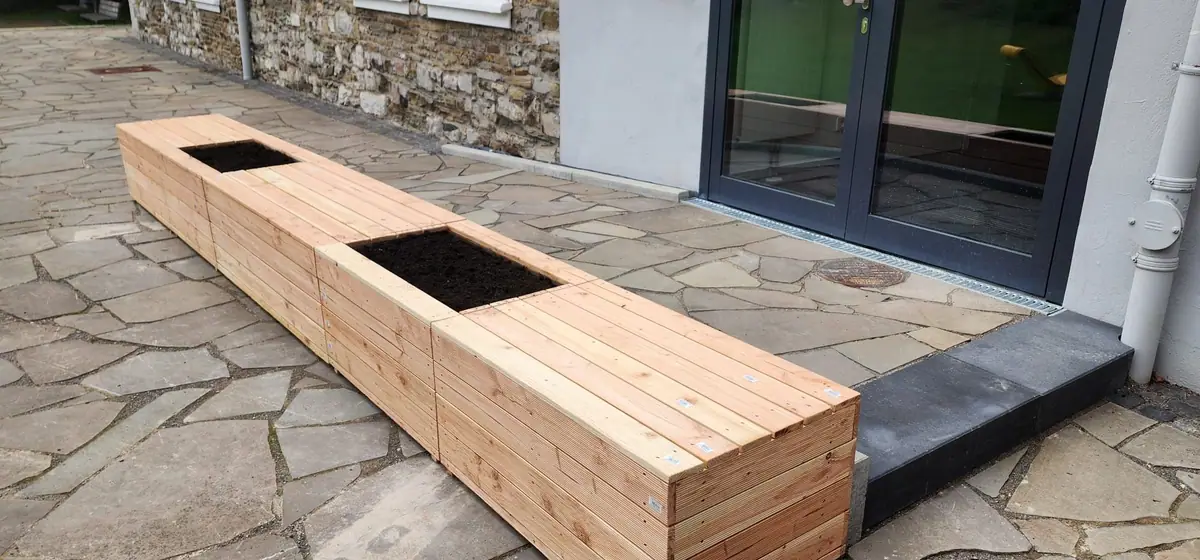 Lange Sitzbank aus Holz mit zwei befüllten Pflanzkästen auf Polygonalplatten, dahinter eine anthrazitfarbene Blockstufe und eine Glastür